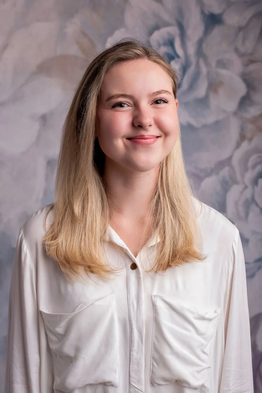 Thalia, head and shoulders, smiling in front of a painted floral backdrop